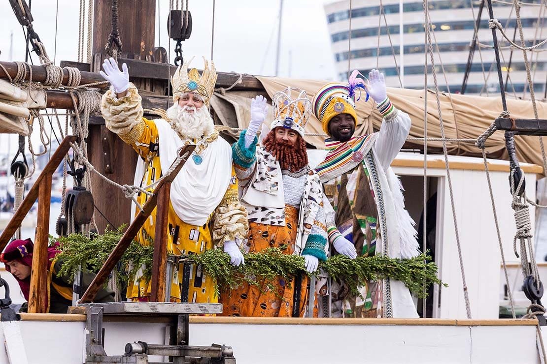 The Three Kings waving from a decorated boat at Port Vell during Barcelona’s Three Kings Parade.