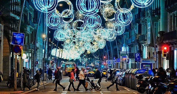 Festive street lights in Barcelona, with people walking through a busy avenue lined with shops during the holiday season.